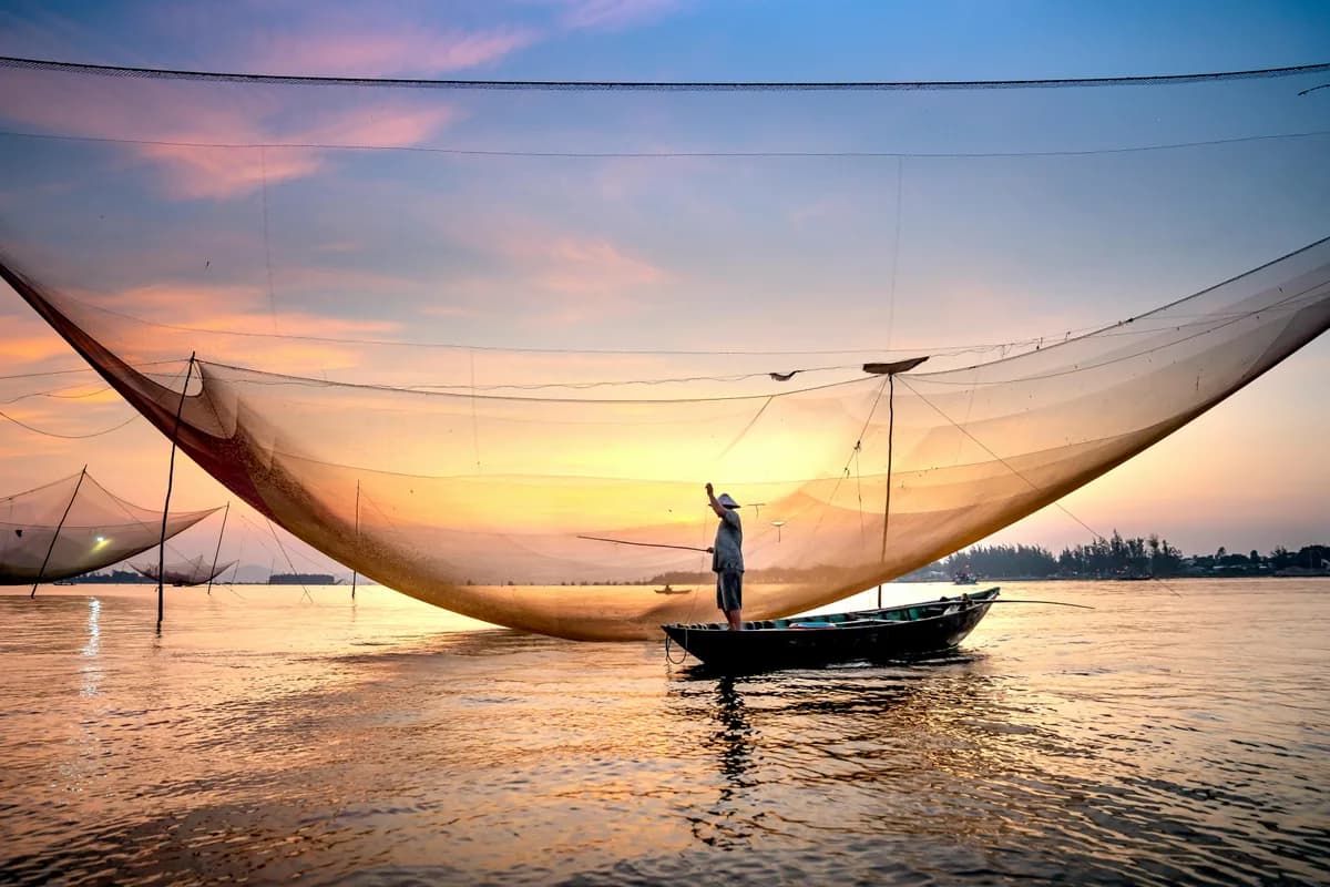 A fisherman casts his net at sunset on the waters of Hội An, Vietnam, showcasing traditional fishing practices.